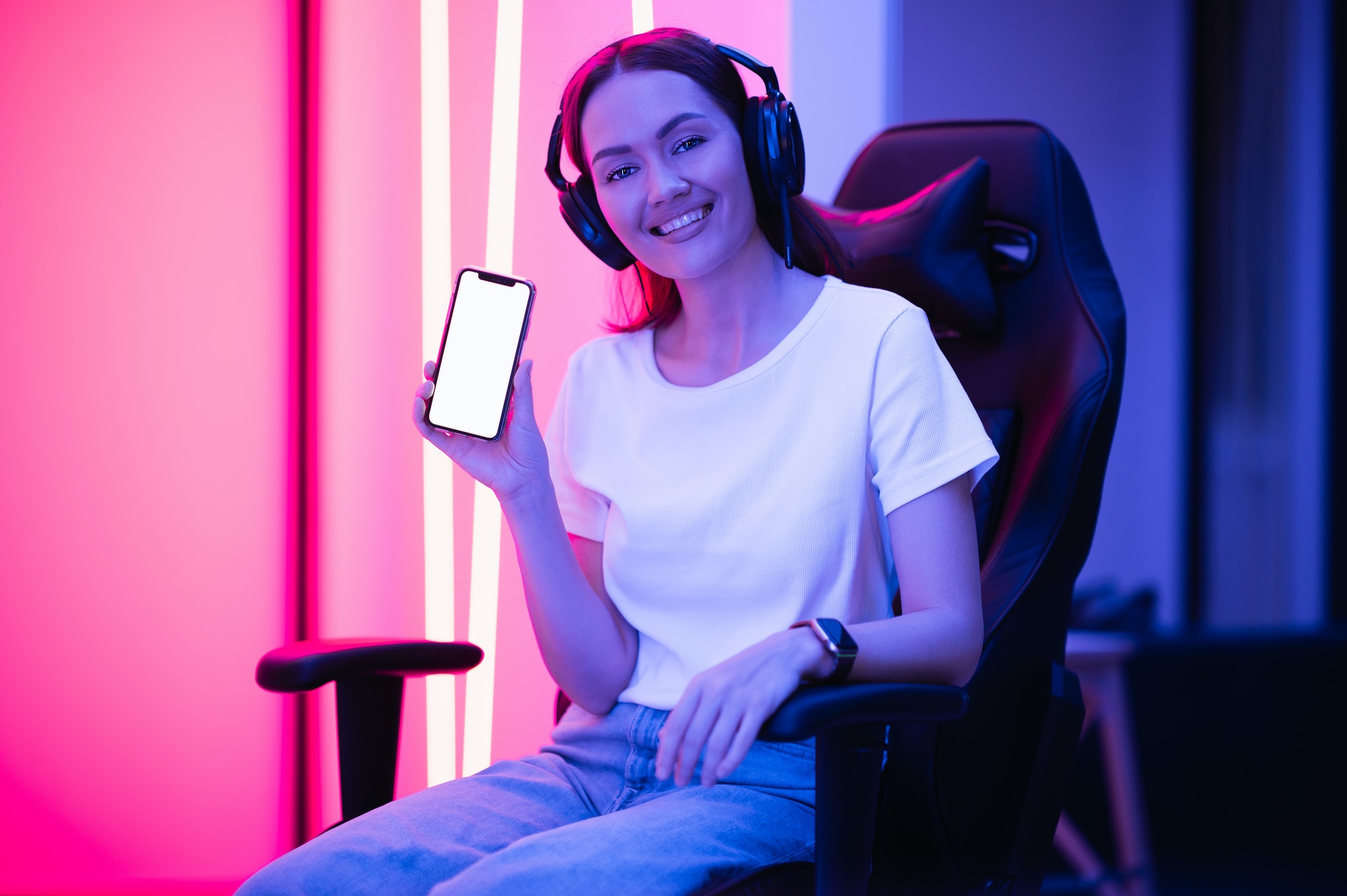 Happy gamer girl wearing headset showing smart phone display and pointing at blank screen with her