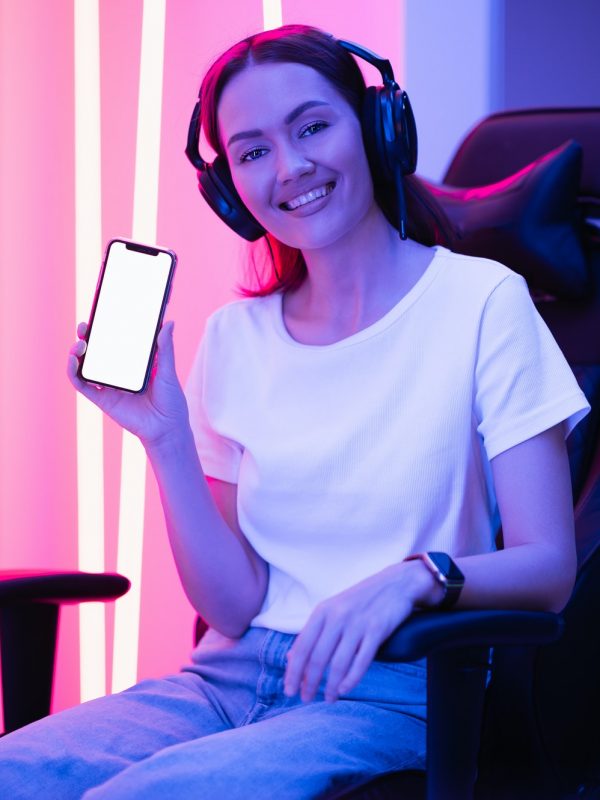 Happy gamer girl wearing headset showing smart phone display and pointing at blank screen with her