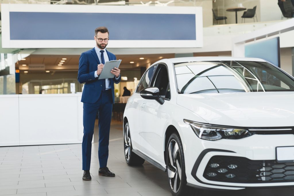 Shop assistant holding clipboard checking car options information at automobile dealer shop store.