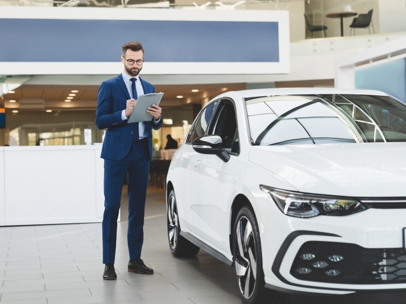 Shop assistant holding clipboard checking car options information at automobile dealer shop store.